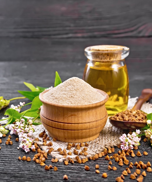 Farina di grano saraceno marrone in una ciotola, semole marroni in un cucchiaio, fiori e foglie di grano saraceno, olio in un barattolo di vetro su tela sullo sfondo di una tavola di legno scuro