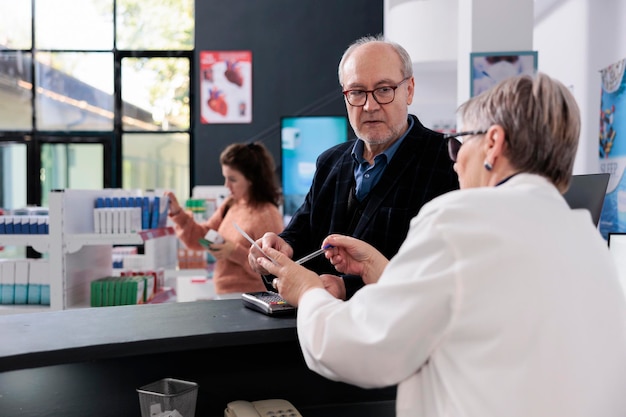 Farmacista anziano in piedi al bancone della farmacia che guarda la prescrizione sanitaria del cliente, aiutandolo con il trattamento farmaceutico che lavora in farmacia. Persone che acquistano vitamine, integratori