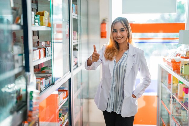 Farmacista attraente donna in uniforme sorridente con il pollice in alto in farmacia