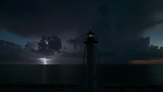 Faro alto bianco sulla riva del mare con luce lampeggiante nella notte tempestosa per la navigazione di navi commerciali Temporale con fulmini sull'acqua dell'oceano che rappresenta un pericolo per le navi