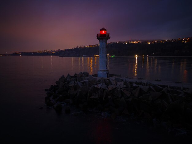 Faro in riva al mare con luce rossa di notte sullo sfondo delle luci delle navi