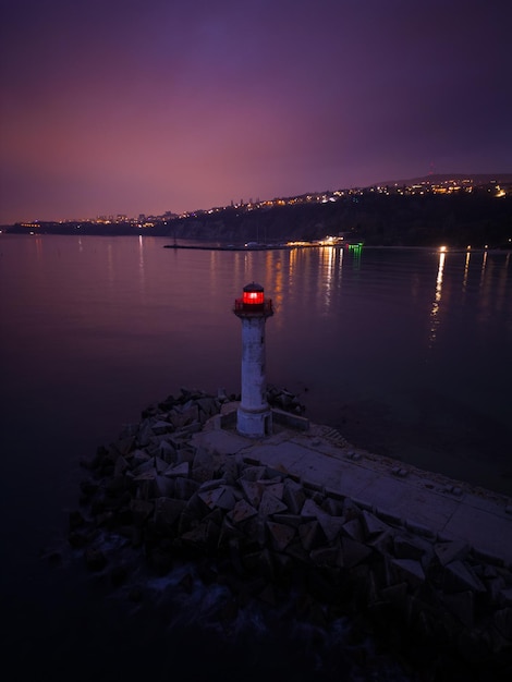 Faro in riva al mare con luce rossa di notte sullo sfondo delle luci delle navi