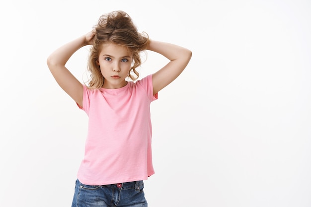 Fascino giovane bambina bionda carina con gli occhi azzurri in maglietta rosa che alza i capelli, facendo una crocchia per capelli, aspetto tenero, macchina fotografica con sguardo alla moda, espressione seria determinata, muro bianco in piedi