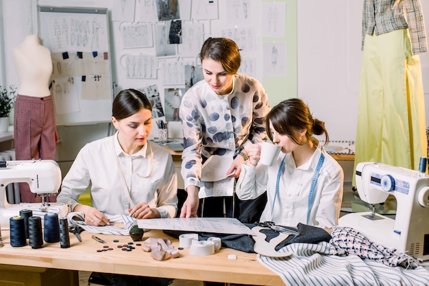Fashion Design, sarta, sarto Concept. Tre giovani donne caucasiche cucitrici che lavorano insieme presso l'atelier luminoso, preparando la nuova collezione di abiti fatti a mano