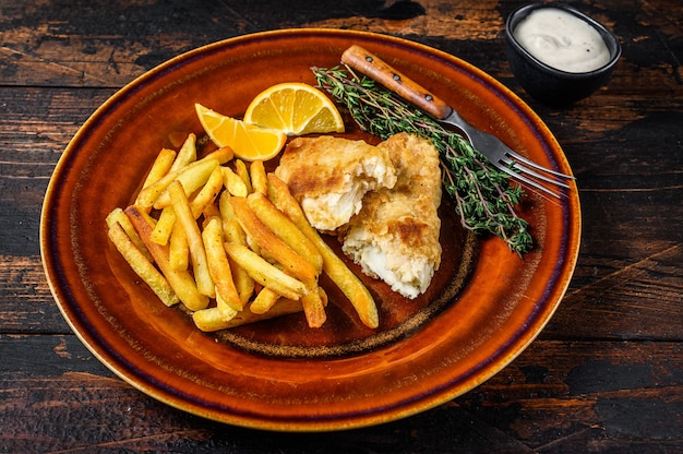 Fast food britannico di pesce e patatine fritte con patatine fritte e salsa tartara su un piatto rustico. Fondo in legno scuro. Vista dall'alto.