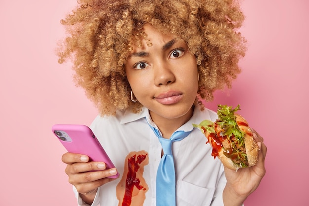 Fast food e cheat meal day Una donna europea seria usa il messaggio sms dei tipi di telefoni cellulari mangia un delizioso hamburger vestito con abiti formali spalmati di ketchup isolato su sfondo rosa