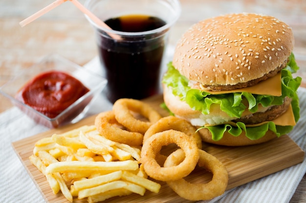 fast food e concetto di mangiare malsano - primo piano di hamburger o cheeseburger, anelli di calamari fritti, patatine fritte, bevanda alla coca cola e ketchup su tavola di legno