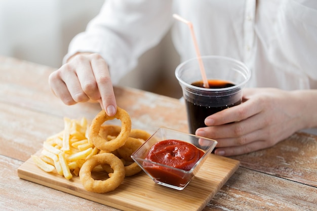 fast food, persone e concetto di alimentazione malsana - primo piano di una donna che mangia anelli di calamari fritti, patatine fritte con ketchup e beve coca cola su un tavolo di legno