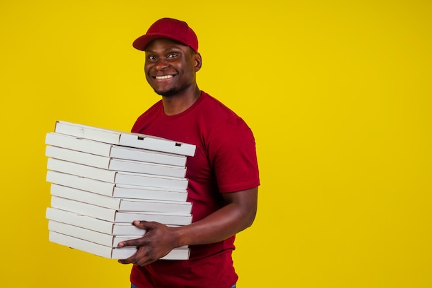 Fattorino afro-americano con pacco che indossa una maglietta rossa e un berretto su sfondo giallo da studio. Concetto di cibo a consegna veloce.