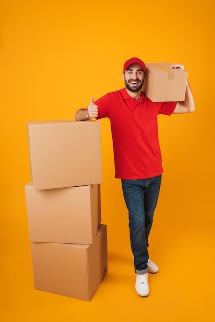 Fattorino caucasico a figura intera in uniforme rossa che sorride mentre sta in piedi con scatole di imballaggio isolate su giallo
