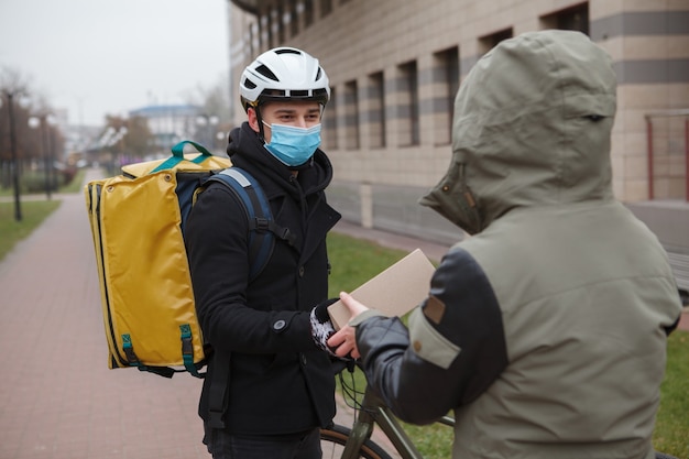Fattorino che indossa una maschera medica che consegna una scatola di cartone a un cliente, che indossa una maschera medica durante la quarantena del coronavirus