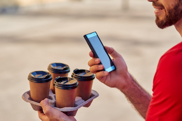 Fattorino che tiene smartphone e tazze di caffè a bordo delle consegne all'aperto, ritagliato. Concetto di consegna del cibo