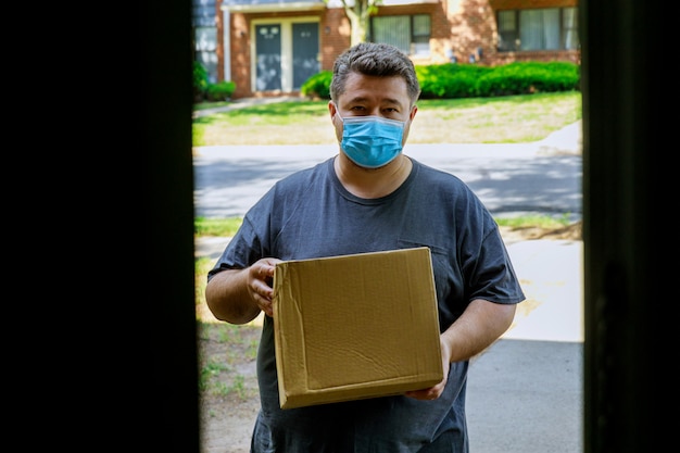 Fattorino con maschera facciale che consegna una scatola di cartone all'ingresso della consegna a domicilio durante la quarantena della pandemia di coronavirus.