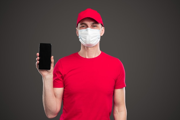 Fattorino in uniforme e maschera che mostra l'app su smartphone con schermo nero mentre pubblicizza il servizio logistico durante l'epidemia di coronavirus su sfondo grigio