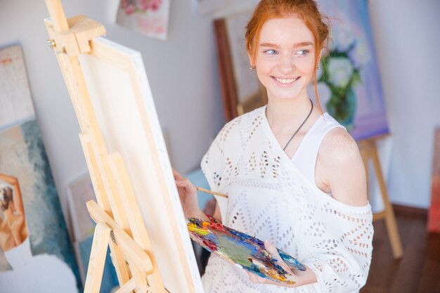 Felice adorabile giovane donna pittore che tiene tavolozza con colori ad olio e pittura su tela in studio d'arte