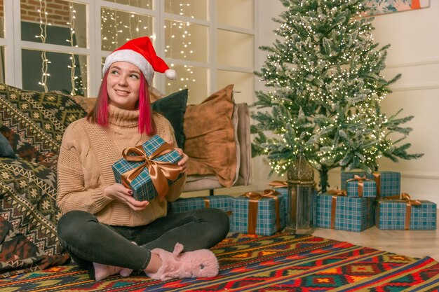 Felice affascinante giovane donna con un maglione con un regalo in mano sul pavimento sullo sfondo di un albero di Natale con regali