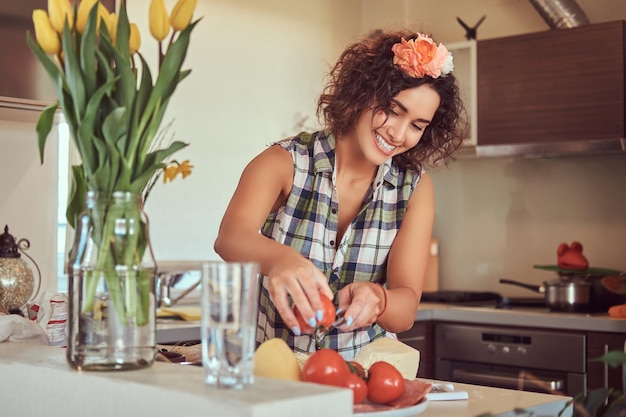 Felice affascinante riccia ragazza ispanica affetta i pomodori mentre cucina nella sua cucina.