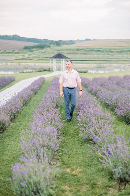 Felice agricoltore maschio maturo che indossa abiti eleganti, camminando con espressioni soddisfatte nella sua bellissima piantagione di lavanda in una giornata estiva