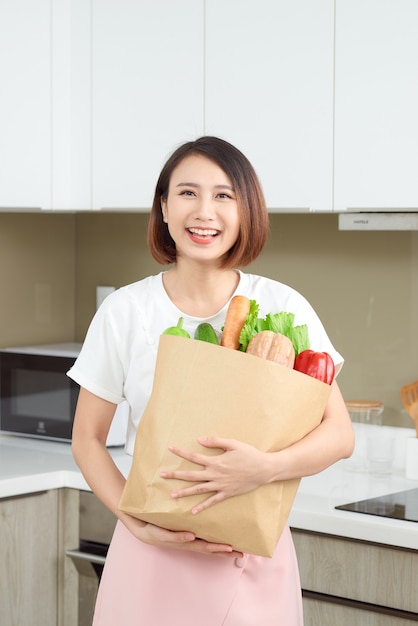 Felice, allegra donna asiatica che tiene in mano un sacchetto di carta pieno di prodotti freschi, verdure, verdure, baguette e banane.