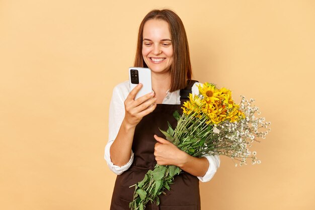 Felice allegro fioraio donna positiva ottimista in piedi con lo smartphone che tiene bouquet di fiori che esprimono la felicità femmina che indossa grembiule marrone