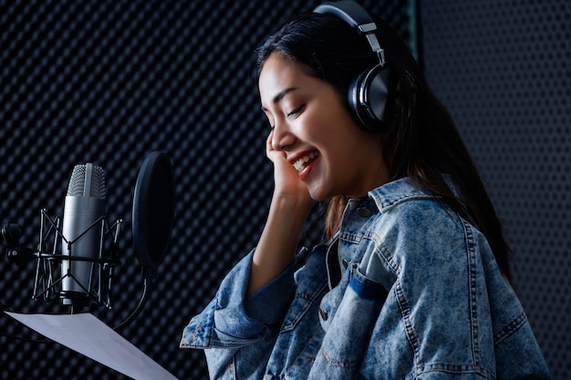 Felice allegro piuttosto sorridente del ritratto di una giovane cantante asiatica che indossa le cuffie che registra una canzone davanti al microfono in uno studio professionale