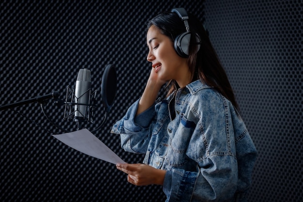 Felice allegro piuttosto sorridente del ritratto di una giovane cantante asiatica che indossa le cuffie che registra una canzone davanti al microfono in uno studio professionale