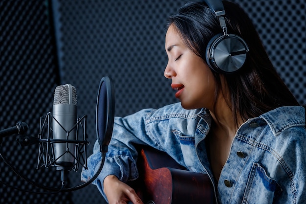 Felice allegro piuttosto sorridente del ritratto di una giovane cantante asiatica che indossa le cuffie con una chitarra che registra una canzone davanti al microfono in uno studio professionale