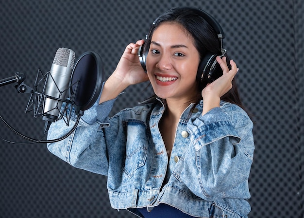 Felice allegro piuttosto sorridente del ritratto di una giovane cantante asiatica che indossa le cuffie con una chitarra che registra una canzone davanti al microfono in uno studio professionale