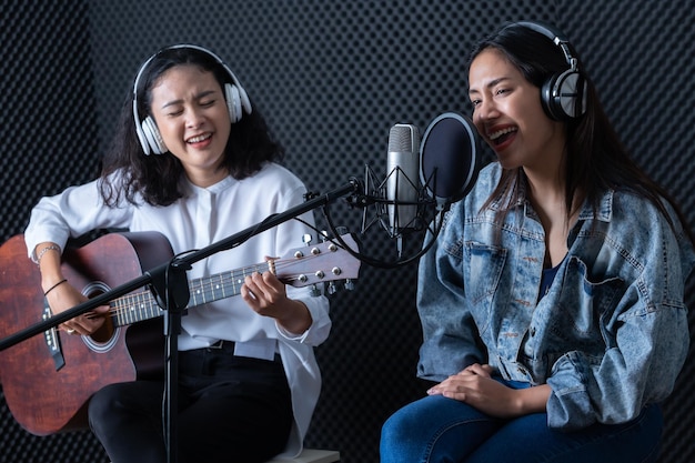 Felice allegro piuttosto sorridente del ritratto Due giovane cantante asiatica donna che indossa le cuffie con una chitarra che registra una canzone davanti al microfono in uno studio professionale