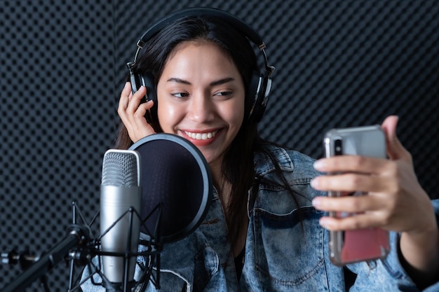 Felice allegro sorridente grazioso del ritratto giovane donna asiatica cantante che indossa le cuffie prendendo un selfie con lo smartphone durante la registrazione di una canzone davanti al microfono in uno studio professionale