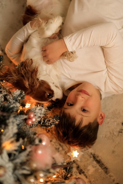 Felice anno nuovo un ragazzo in pigiama leggero fatto in casa abbraccia il suo animale domestico cavalier king charles spaniel a casa nella camera da letto vicino all'albero di Natale