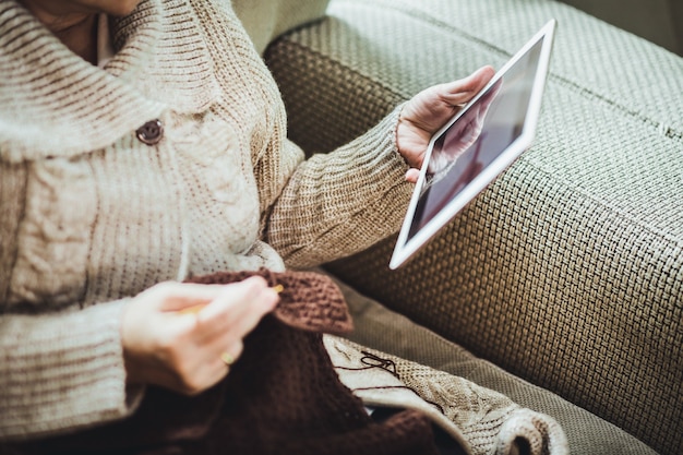 Felice anziana asiatica lavora a maglia sciarpa marrone all'uncinetto e tablet sul divano nel soggiorno di casa