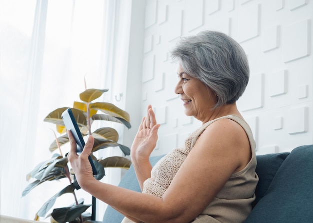 Felice asiatico senior casual donna seduta sul divano salutando lo schermo del tablet digitale in mano nella stanza bianca Le donne anziane si incontrano con la famiglia tramite videochiamata a casa Le persone anziane con la tecnologia