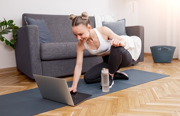 Felice atleta femminile in abbigliamento sportivo seduto sulla stuoia vicino al divano e navigando sul laptop durante l'allenamento durante l'auto isolamento a casa