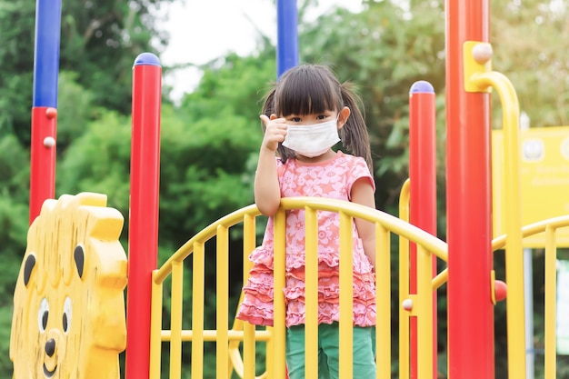 Felice bambina asiatica che sorride e indossa una maschera facciale che mostra il pollice in su le dita