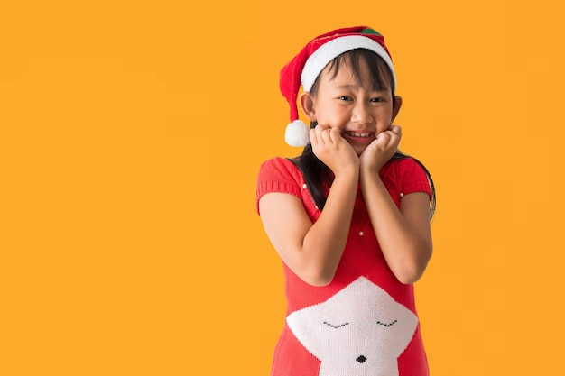 Felice bambina asiatica sorridente con il cappello di Babbo Natale con un costume rosso di Natale che sembra sorpresa posando isolata su uno sfondo giallo