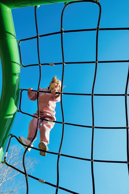 Felice bambina attiva si arrampicò su una rete di corda in un parco giochi contro un cielo blu brillante