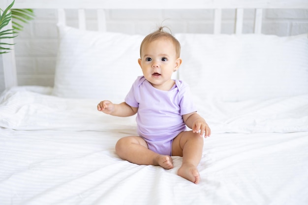 Felice bambina carina in tuta si siede sul letto su biancheria da letto bianca, sorridente. Il concetto di prodotti per bambini.