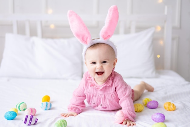 Felice bambina caucasica con un bordo di orecchie da coniglio sulla testa giace sul letto a casa in camera da letto con colorate uova di Pasqua pasqua bambino