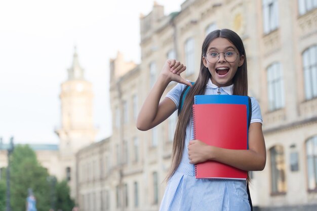 Felice bambina con gli occhiali che puntano il dito sui libri scolastici all'aperto, informazioni, spazio per copie.