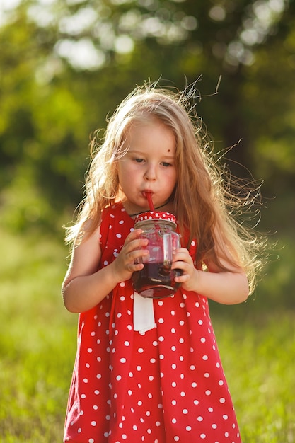 Felice bambina di 3 anni in abito rosso sta bevendo succo nel giardino estivo del villaggio Godimento della natura