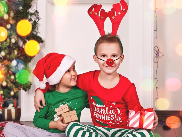 Felice bambina e ragazzo sorridenti con berretto da Babbo Natale e maschera di cervo seduti sulla coperta in pigiama natalizio con confezione regalo.