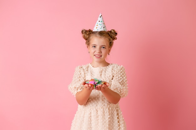 Felice bambina in un berretto di compleanno soffia via i suoi palmi coriandoli multicolori su uno sfondo rosa in studio