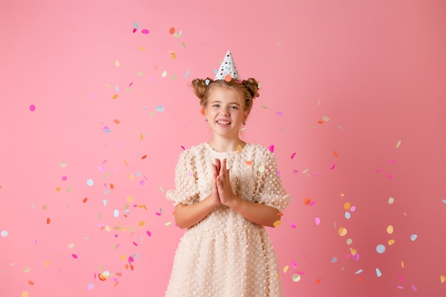 Felice bambina in un berretto di compleanno soffia via i suoi palmi coriandoli multicolori su uno sfondo rosa in studio