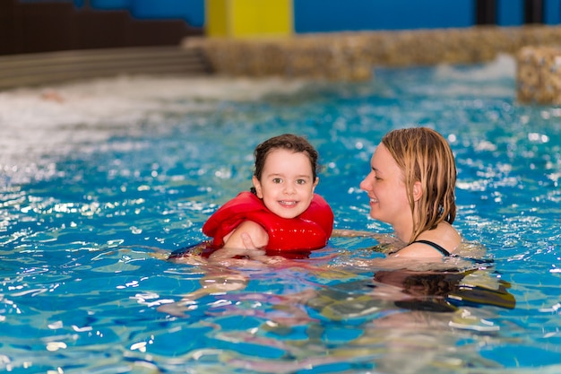 felice bambina in un giubbotto rosso sta nuotando con sua madre nella piscina del parco acquatico