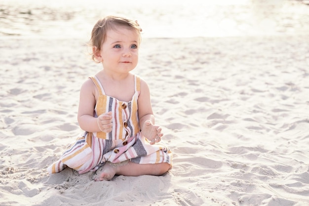 felice bambina seduta sulla spiaggia sulla sabbia che indossa un abito estivo a righe in una giornata di sole