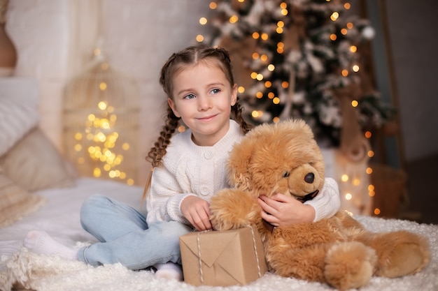 Felice bambina sorridente e gioca con un grande orsacchiotto e apre il regalo di Natale a casa