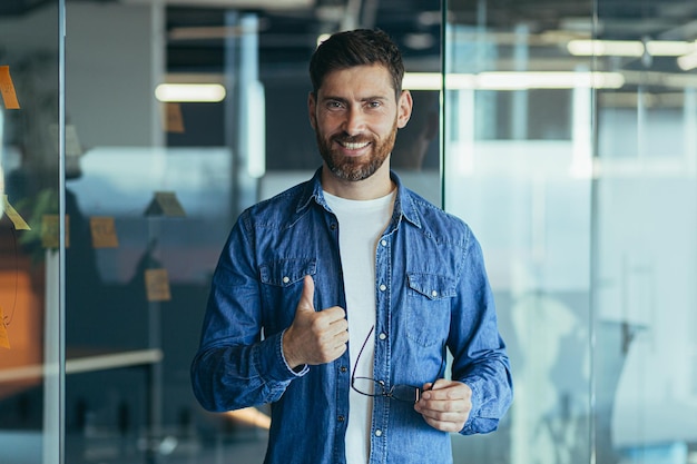 Felice barbuto imprenditore proprietario di avvio in piedi in un ufficio moderno guardando la fotocamera sorridente giovane uomo creativo hipster in posa nel ritratto di affari dell'area di lavoro