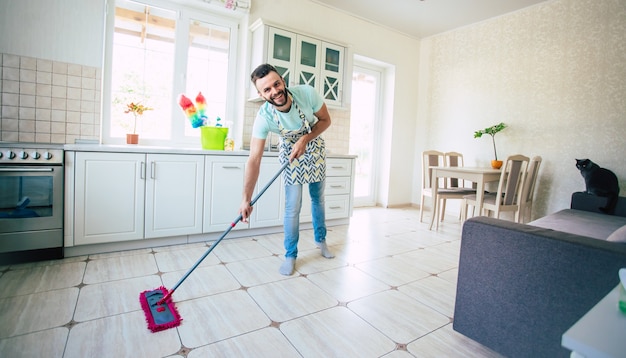 Felice bel giovane barba uomo sta pulendo il pavimento nella cucina domestica e divertirsi.
