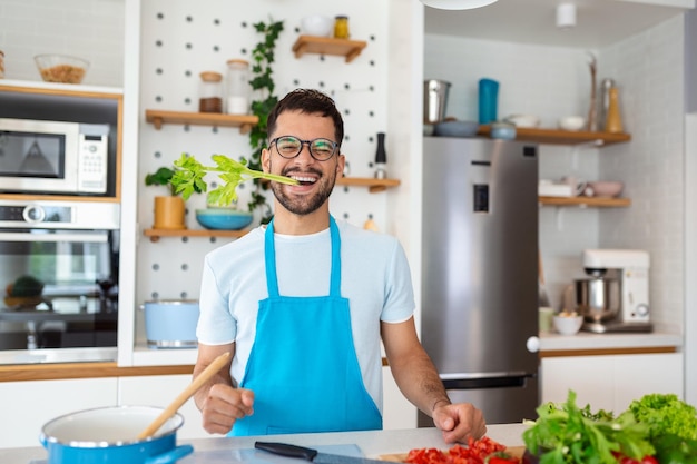 Felice bell'uomo che cucina in cucina a casa con il sedano in bocca che posa per la foto Concetto di vita sana